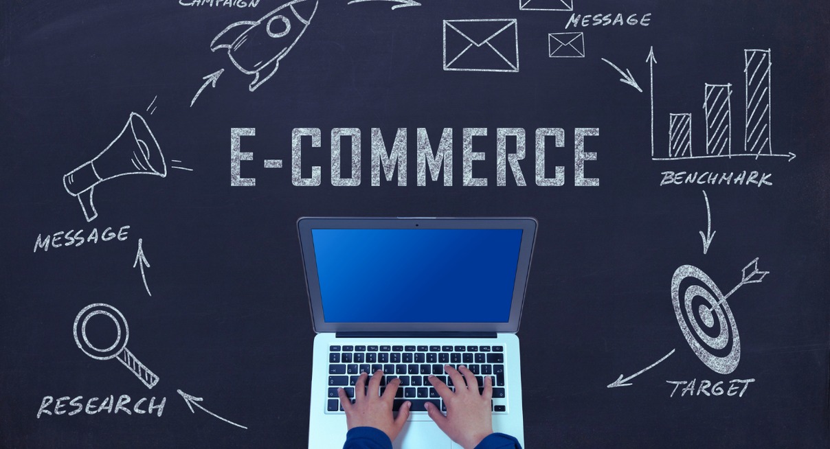 Mains sur un laptop devant un tableau noir avec le mot E-COMMERCE entouré de concepts de stratégie digitale