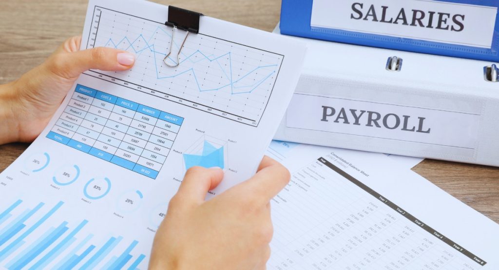Mains féminines tenant des rapports de coûts salariaux avec des classeurs Salaries et Payroll en arrière-plan